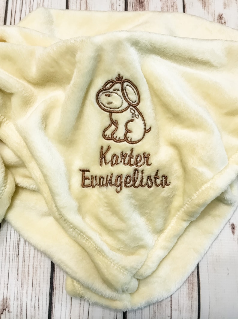 Pode incluir: Um cobertor de pel&uacute;cia de cor creme com um cachorro bordado em marrom e o nome "Karter Evangelista" bordado abaixo.