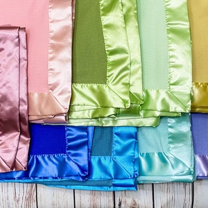 May include: A collection of ten baby blankets with satin trim in various colors, including pink, green, blue, purple, and gray. The blankets are arranged in a row, with the satin trim facing the viewer.
