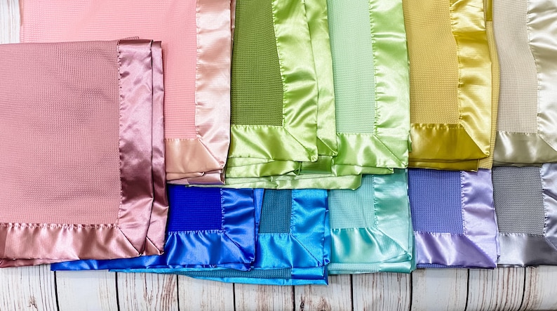 May include: A collection of ten baby blankets with satin trim in various colors, including pink, green, blue, purple, and gray. The blankets are arranged in a row, with the satin trim facing the viewer.