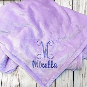 Pode incluir: Um cobertor macio de pel&uacute;cia roxo com um monograma "M" e o nome "Mirella" bordado em fio azul.