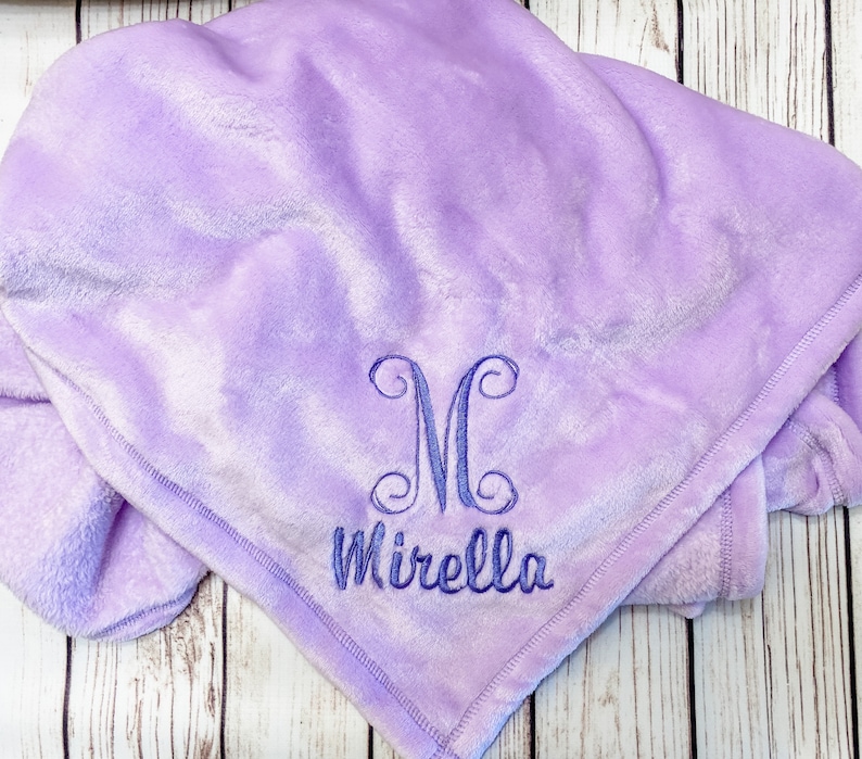 Pode incluir: Um cobertor macio de pel&uacute;cia roxo com um monograma "M" e o nome "Mirella" bordado em fio azul.