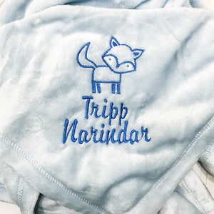 Pode incluir: Um cobertor de beb&ecirc; azul claro com uma raposa bordada em azul e o nome "Tripp Narindar".