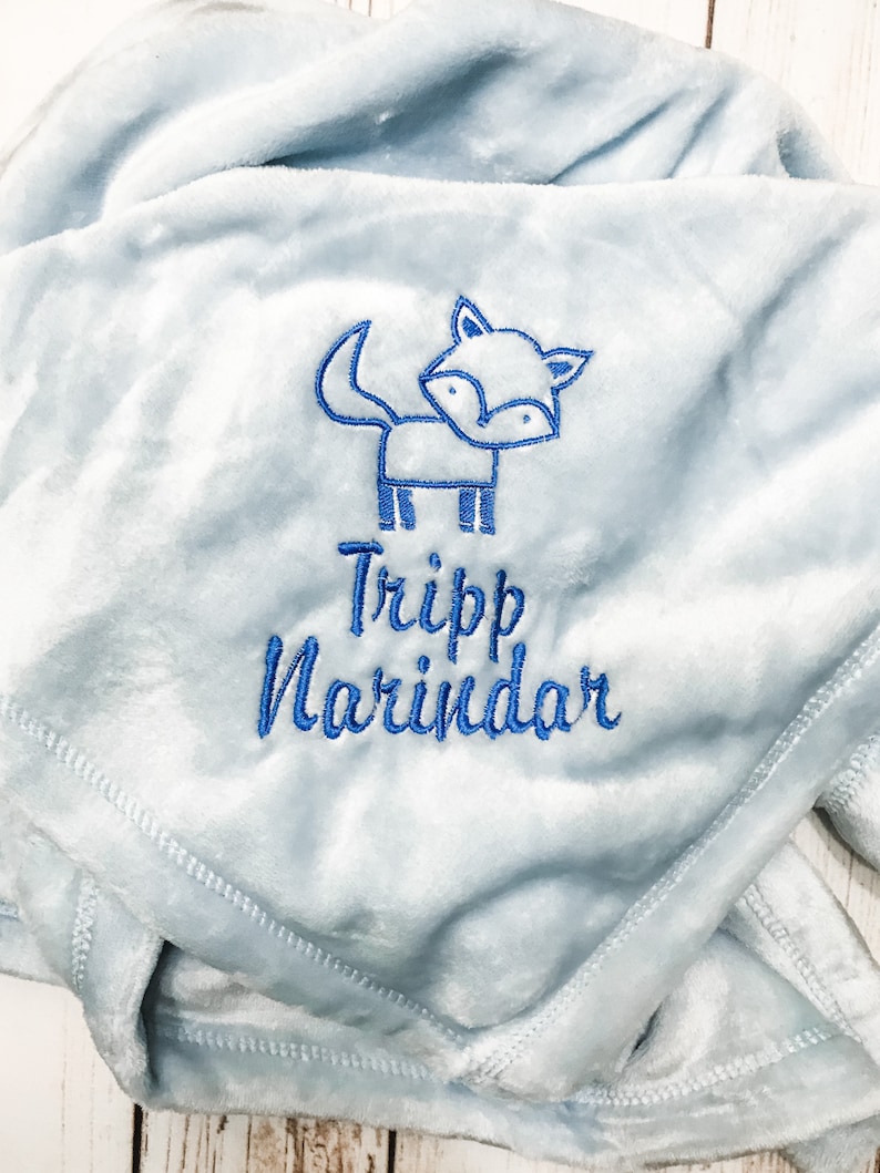 Pode incluir: Um cobertor de beb&ecirc; azul claro com uma raposa bordada em azul e o nome "Tripp Narindar".