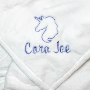 Pode incluir: Cobertor de pel&uacute;cia branco com um unic&oacute;rnio bordado em azul e o nome "Cora Joe"
