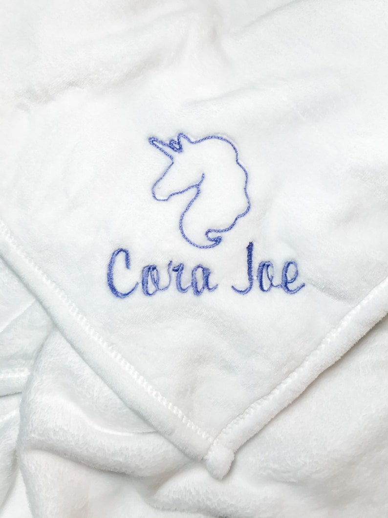 Pode incluir: Cobertor de pel&uacute;cia branco com um unic&oacute;rnio bordado em azul e o nome "Cora Joe"
