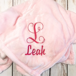 Pode incluir: Um cobertor de pel&uacute;cia rosa com o nome "Leah" bordado em letras cursivas rosa.