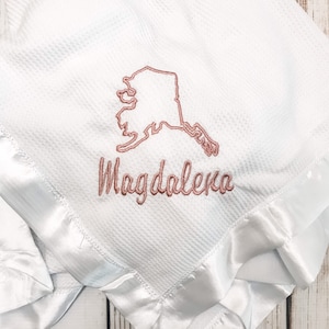 May include: White waffle weave baby blanket with a satin trim. The blanket is embroidered with the outline of the state of Alaska and the name "Magdalena".