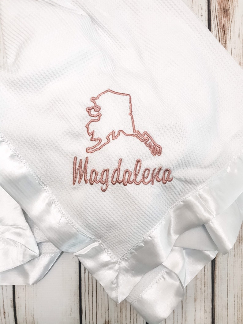May include: White waffle weave baby blanket with a satin trim. The blanket is embroidered with the outline of the state of Alaska and the name "Magdalena".