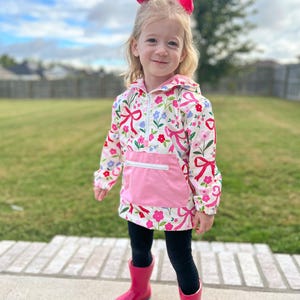 May include: A young child wearing a white floral print jacket with pink bows and flowers, a pink pocket, black leggings, and bright pink rain boots. A large pink bow adorns the child's hair. The background features a green lawn and a cloudy sky.