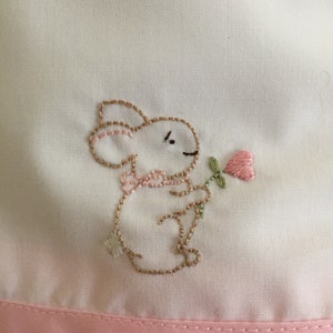 May include: A white fabric with a pink embroidered bunny holding a pink flower. The fabric has a pink trim around the edge.