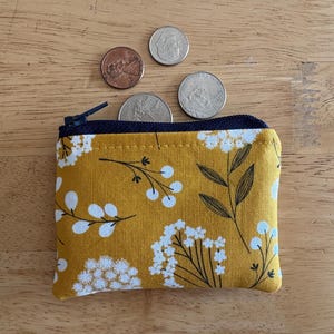 May include: A small mustard yellow zippered pouch with a white floral pattern. The pouch is lying on a wooden surface with four coins scattered around it.