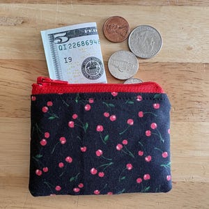 May include: A small, black fabric wallet with a red zipper and a cherry pattern. A five-dollar bill and several coins are visible. The wallet is open, revealing the red interior.