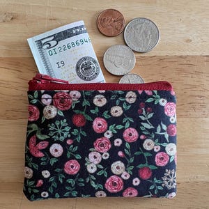 May include: A small, black floral print wallet with a red zipper, containing a five-dollar bill and several coins. The wallet features a pattern of pink and red roses with green leaves.