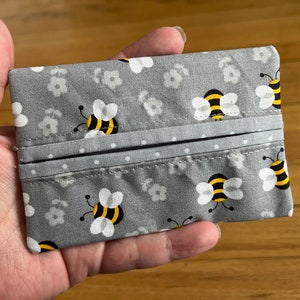 Puede incluir: Un porta pañuelos de tela gris con un patrón de abejas y flores. El porta pañuelos tiene una solapa que se pliega para cerrarse.