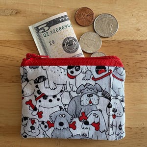 May include: A small, rectangular coin purse with a red zipper and trim, filled with a five-dollar bill. The purse is patterned with cartoon dogs in various poses, holding bones. Several coins are scattered around the purse on a wooden surface.