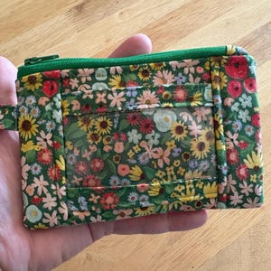 Peut inclure: Une pochette zippée verte avec une fenêtre en plastique transparent. La pochette est décorée d'un imprimé floral avec des fleurs roses, rouges et jaunes sur un fond vert.