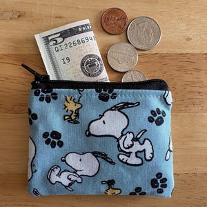 May include: A small, light blue zippered coin purse with a Snoopy and Woodstock pattern, and paw prints. A five-dollar bill and several coins are next to the purse on a wooden surface.