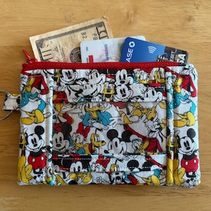 Peut inclure: Une pochette zippée rouge et blanche avec un imprimé Mickey Mouse et ses amis. La pochette a un porte-clés et est remplie de cartes et d'argent liquide.
