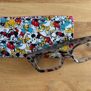 May include: A pair of eyeglasses with a tortoiseshell frame rests on a wooden surface next to a rectangular glasses case. The case is covered in a colorful pattern featuring Mickey Mouse, Minnie Mouse, and Donald Duck.