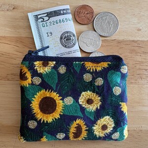 May include: A small, zippered coin purse with a sunflower pattern, containing a five-dollar bill and several coins. The purse is navy blue with yellow sunflowers, green leaves, and gold accents. The coins include a penny, nickel, and quarter.