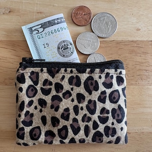 May include: A small, leopard print coin purse with a black zipper, partially open to reveal a five-dollar bill. Several coins, including a penny, a nickel, a dime, and a quarter, are scattered around the purse on a wooden surface.