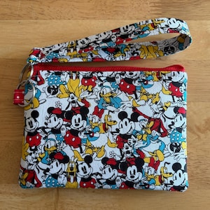 Peut inclure: Un porte-monnaie rectangulaire blanc avec une fermeture éclair rouge et une dragonne. Le sac est recouvert d'un motif coloré de Mickey Mouse, Minnie Mouse, Donald Duck et d'autres personnages Disney. La dragonne a le même motif.