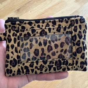 May include: A rectangular leopard print wallet with a clear ID window and a black zipper closure. The wallet has a key ring attached to the side. The wallet is held in a hand.