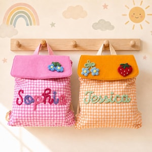 Könnte beinhalten: Zwei Stoffrucksäcke mit Vichy-Karo-Muster. Der linke Rucksack ist rosa und mit dem Namen Sophia in Lila bestickt. Der rechte Rucksack ist orange und mit dem Namen Jessica in Grün bestickt. Beide haben gehäkelte Blumen- und Fruchtapplikationen.