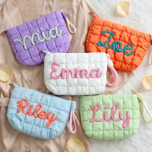 Op de afbeelding: Vijf gewatteerde make-up tasjes in verschillende kleuren: paars, oranje, wit, blauw en groen. Elke tas is geborduurd met een andere naam in een contrasterende kleur. De tasjes hebben een polsbandje en een ritssluiting.