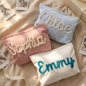 Puede incluir: Cuatro neceseres de pana en colores pastel: rosa, azul claro y blanco. Cada neceser presenta un nombre bordado en un color contrastante: "Sophia", "Chloe" y "Emmy". Los neceseres están colocados sobre una superficie suave de tonos neutros.