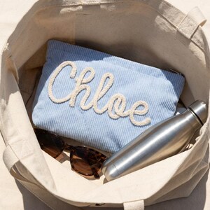 May include: A beige canvas tote bag filled with a light blue corduroy pouch with the name "Chloe" in white rope lettering, sunglasses, and a silver water bottle.