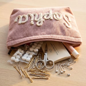 May include: A pink corduroy cosmetic bag with the name "Sophia" embroidered on the front. The bag is open, revealing various items, including bobby pins, safety pins, scissors, and blotting papers.