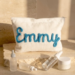 May include: A white corduroy cosmetic bag with the name "Emmy" embroidered in blue yarn. The bag is rectangular with a zipper closure. Several skincare products, including bottles and jars, are arranged in front of the bag.