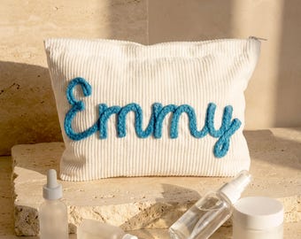 May include: A white corduroy cosmetic bag with the name "Emmy" embroidered in blue yarn. The bag is rectangular with a zipper closure. Several skincare products, including bottles and jars, are arranged in front of the bag.