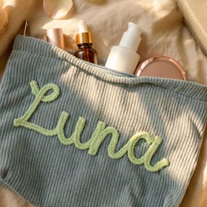 May include: A light blue corduroy cosmetic bag with the name "Luna" in light green stitched letters. The bag is open, revealing several beauty products, including a brown glass bottle with a dropper, a white pump bottle, and a compact.
