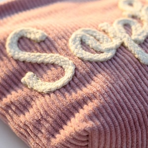 May include: Close-up of a pink corduroy fabric with the word "Soep" embroidered in white, textured yarn. The letters are in a cursive style, adding a handcrafted feel to the textile. The fabric has a ribbed texture.