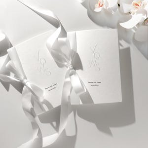 Könnte beinhalten: Zwei weiße Hochzeitsgelübde-Broschüren, gebunden mit weißen Satinbändern. Der geprägte Text "Vows" ist auf der Vorderseite zu sehen, zusammen mit den Namen "Mason and Emma" und dem Datum "08.05.2025".