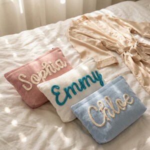 May include: Four corduroy cosmetic bags in pink, white, and blue, each embroidered with a name: Sophia, Emmy, and Chloe. A beige satin robe rests on the bed in the background. The bags are rectangular with a zipper closure.