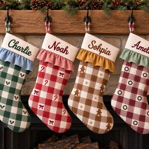 Puede incluir: Cuatro calcetines navideños personalizados colgados en fila. Cada uno tiene un puño color crema con un nombre bordado en rojo o verde oscuro, un borde con volantes y un patrón de cuadros con pequeños iconos festivos como acebo y coronas.