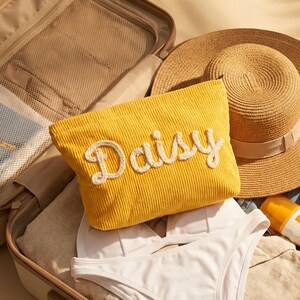 May include: A yellow corduroy travel pouch with the name "Daisy" embroidered in white. The pouch is inside an open suitcase, alongside a straw sun hat and white bikini. A bottle of sunscreen is also visible.