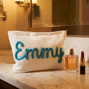 May include: Cream-colored corduroy cosmetic bag with the name "Emmy" embroidered in turquoise. Beside the bag are a perfume bottle and a red lipstick tube, all resting on a marble countertop.