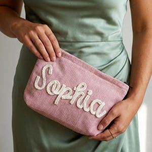 May include: A pink corduroy clutch with the name "Sophia" embroidered in white, held against a green background. The clutch has a zipper closure and a rectangular shape. The corduroy texture is visible.