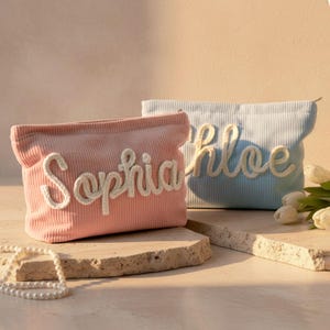 May include: Two corduroy cosmetic bags, one pink and one light blue, each with a name embroidered in a cursive font. The pink bag says "Sophia" and the blue bag says "Chloe". Both bags have a zipper closure.