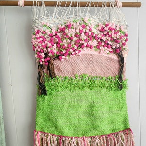 May include: A pink and green woven wall hanging with a floral design. The hanging features a pink background with two trees with pink flowers and green leaves. The bottom of the hanging is green and has pink fringe.