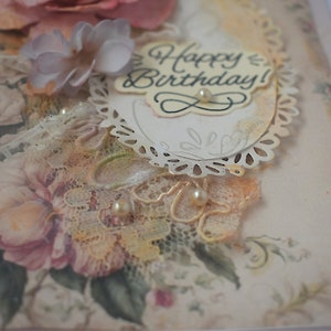 Card is flat on white background. Base is pink shimmer under vintage rose pattern. Left side has handmade & prima flowers in pinks and hand-dyed lace snippet.  Vintage oval frame is in middle showing vintage effect. Sentiment is Happy Birthday.