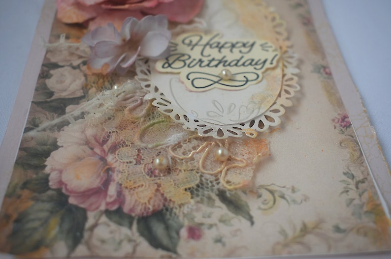 Card is flat on white background. Base is pink shimmer under vintage rose pattern. Left side has handmade & prima flowers in pinks and hand-dyed lace snippet.  Vintage oval frame is in middle showing vintage effect. Sentiment is Happy Birthday.