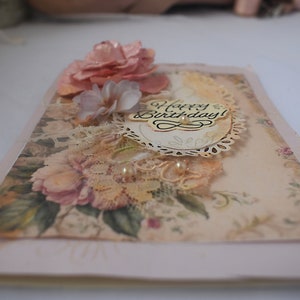 Card is flat on white background. Base is pink shimmer under vintage rose pattern. Left side has handmade & prima flowers in pinks and hand-dyed lace snippet.  Vintage oval frame is in middle showing vintage effect. Sentiment is Happy Birthday.