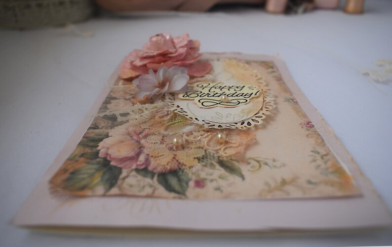 Card is flat on white background. Base is pink shimmer under vintage rose pattern. Left side has handmade & prima flowers in pinks and hand-dyed lace snippet.  Vintage oval frame is in middle showing vintage effect. Sentiment is Happy Birthday.