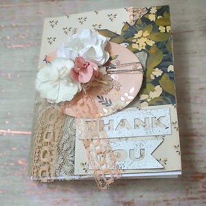 May include: A handmade greeting card with a floral design. The card features a cream-colored background with a small floral pattern, accented with lace, paper flowers, and the words "THANK YOU".