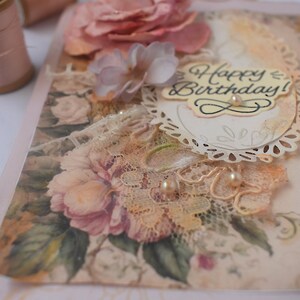 Card is flat on white background. Base is pink shimmer under vintage rose pattern. Left side has handmade & prima flowers in pinks and hand-dyed lace snippet.  Vintage oval frame is in middle showing vintage effect. Sentiment is Happy Birthday.